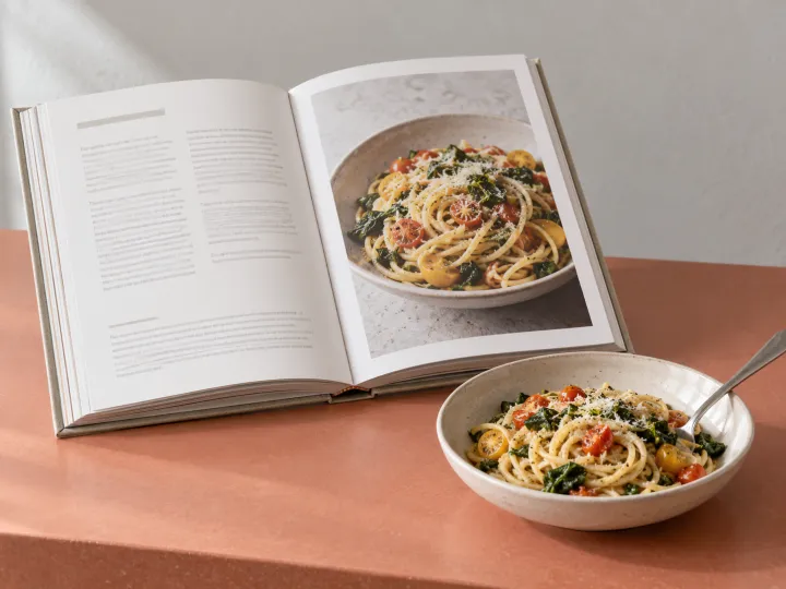 An open hardback cookbook with indistinct recipe text and a pasta photograph beside a matching dish on a coral kitchen counter.