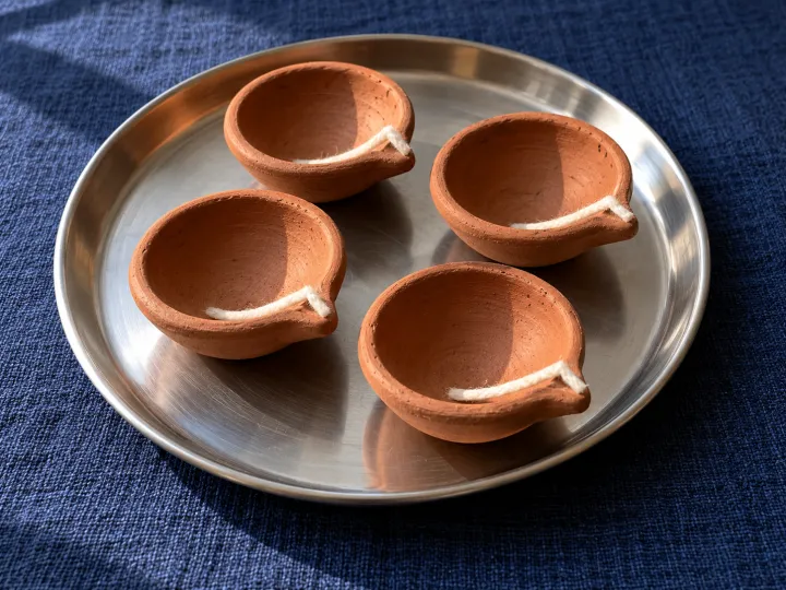 Four unlit terracotta diya lamps with cotton wicks on a metal tray over dark blue fabric.