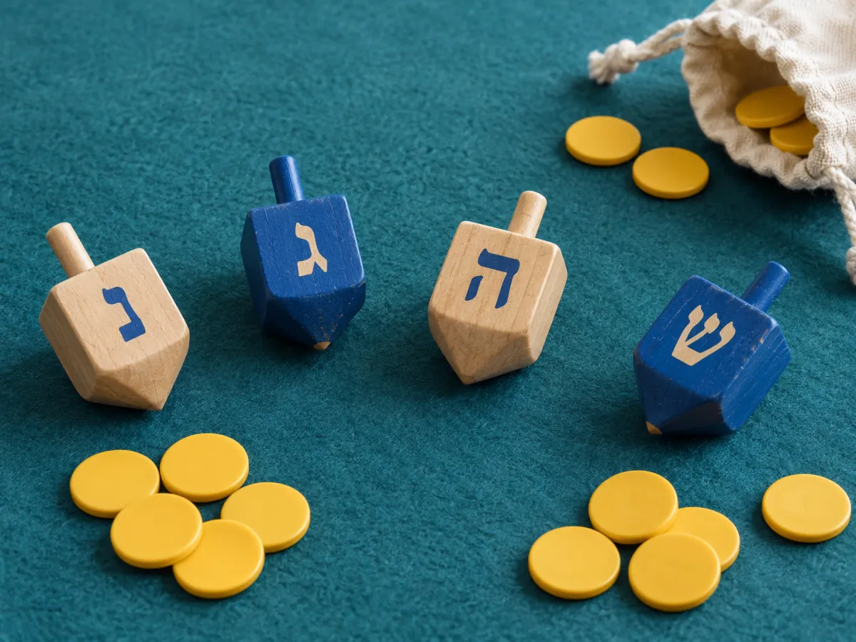 Four wooden dreidels with Hebrew game letters beside yellow counting discs and a small cloth pouch on a teal mat.