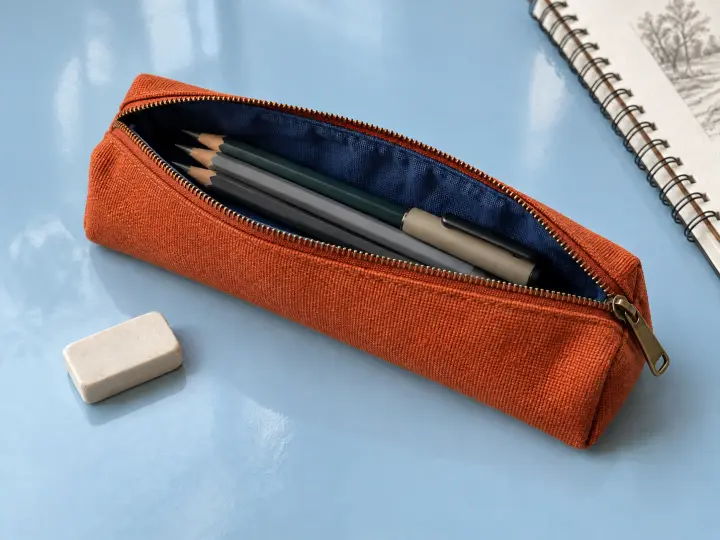 An open rust-orange canvas pencil case holding pencils and a capped pen on a pale blue surface, with an eraser beside it.