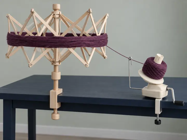 A wooden umbrella swift holding plum yarn beside a cream hand-cranked yarn winder clamped to a navy table.