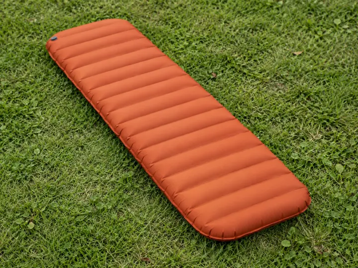 A rust-orange inflatable camping sleeping mat with transverse air chambers and a closed valve on short grass.