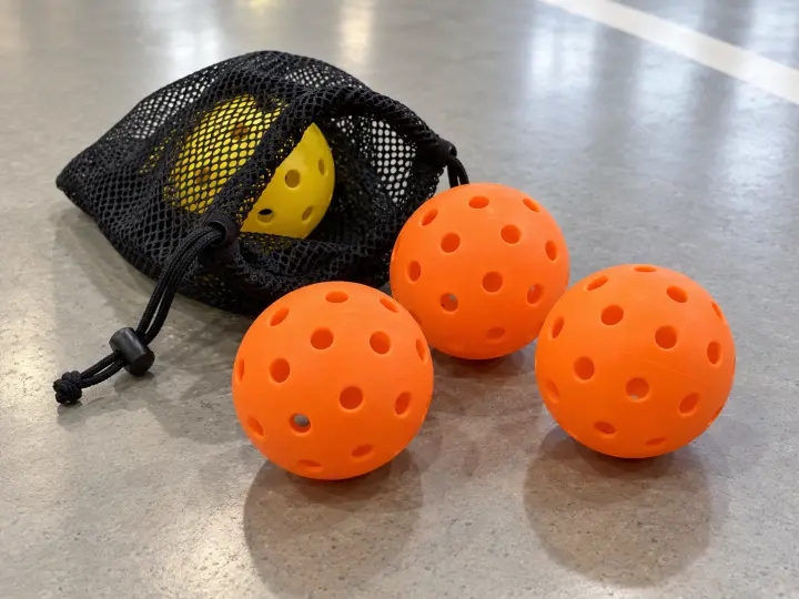 Orange perforated pickleballs beside an open mesh pouch with a yellow ball