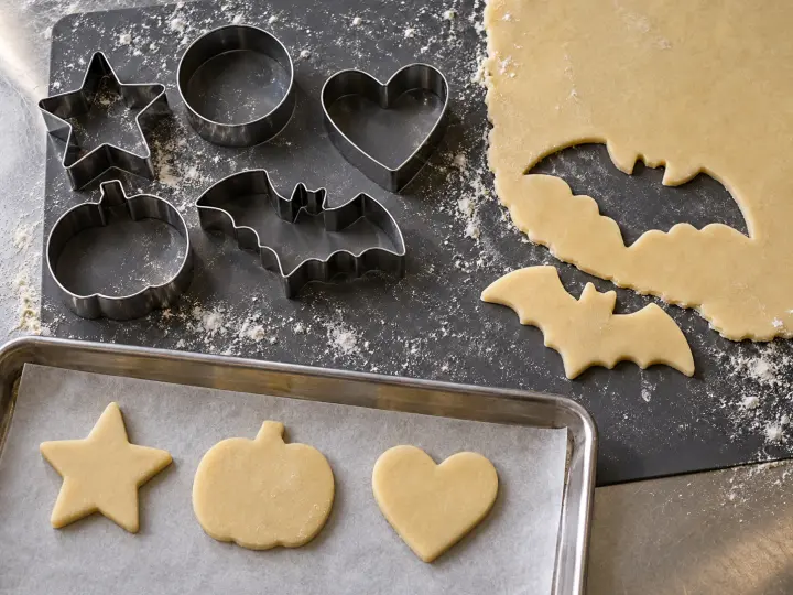 Five hollow metal cookie cutters beside rolled dough with a bat-shaped opening and raw cookies on a tray.