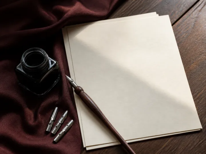 A wooden calligraphy pen, three metal nibs and an ink bottle beside blank cream paper.