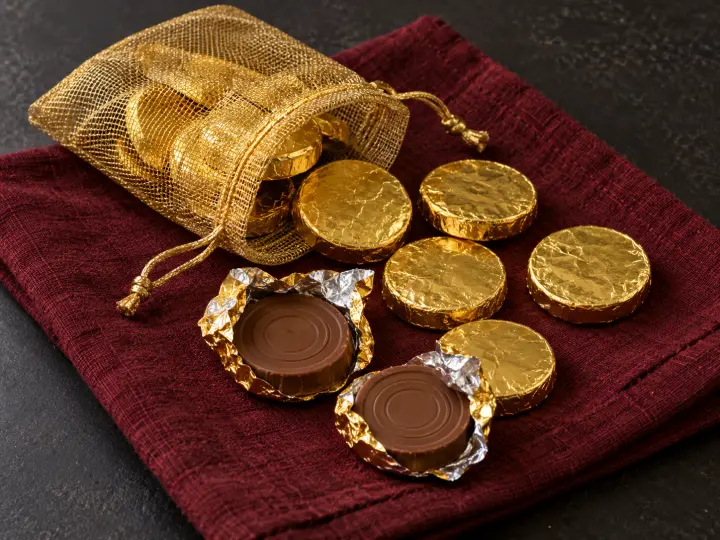 An open gold mesh pouch of foil-wrapped chocolate coins, with two unwrapped chocolate discs on burgundy fabric.