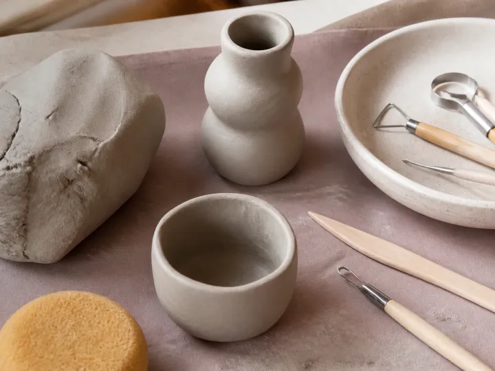 Grey air-dry clay, sculpting tools and two unfinished handmade pots on a protected table.