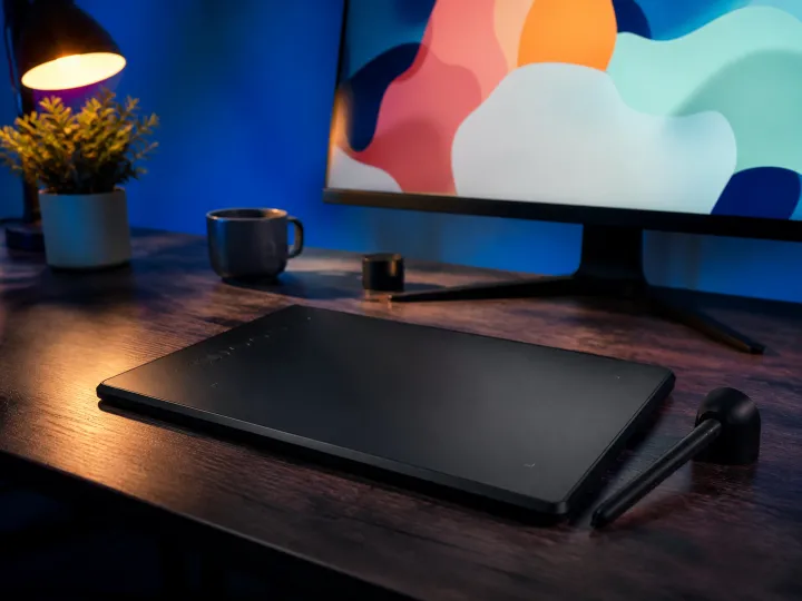 A black pen drawing tablet and stylus on a desk below a monitor showing colourful abstract shapes.