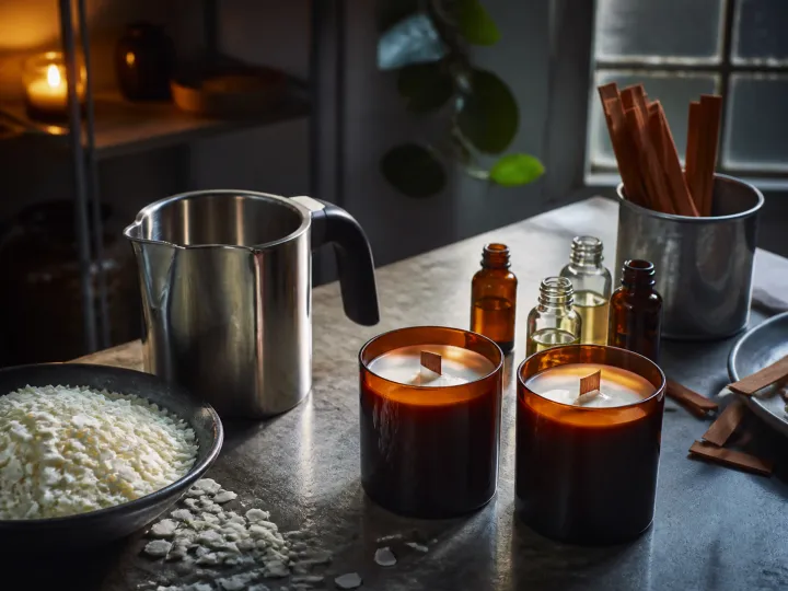 Amber glass candles, wax flakes, fragrance bottles and a metal pouring jug on a work surface.