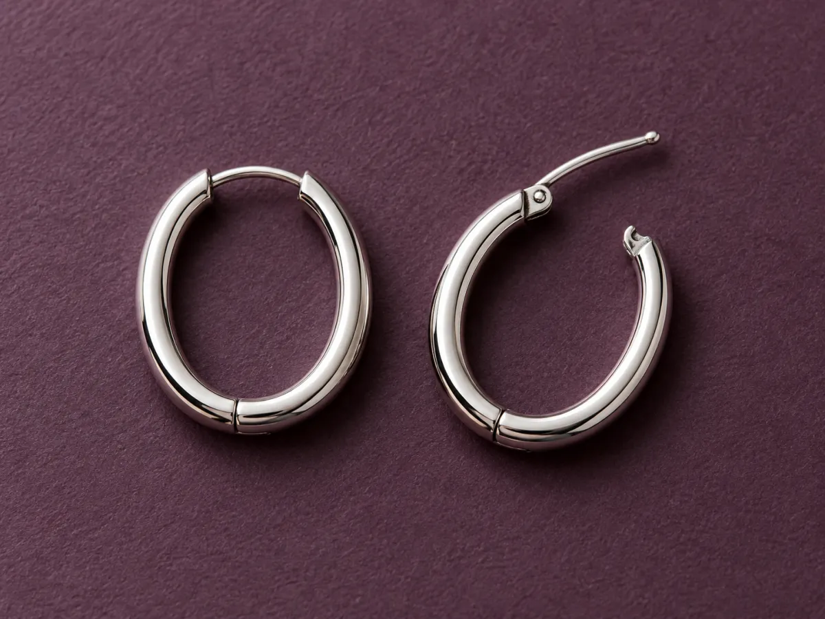 Two polished silver-colored oval hoop earrings on plum paper, with one hinged piercing post open.