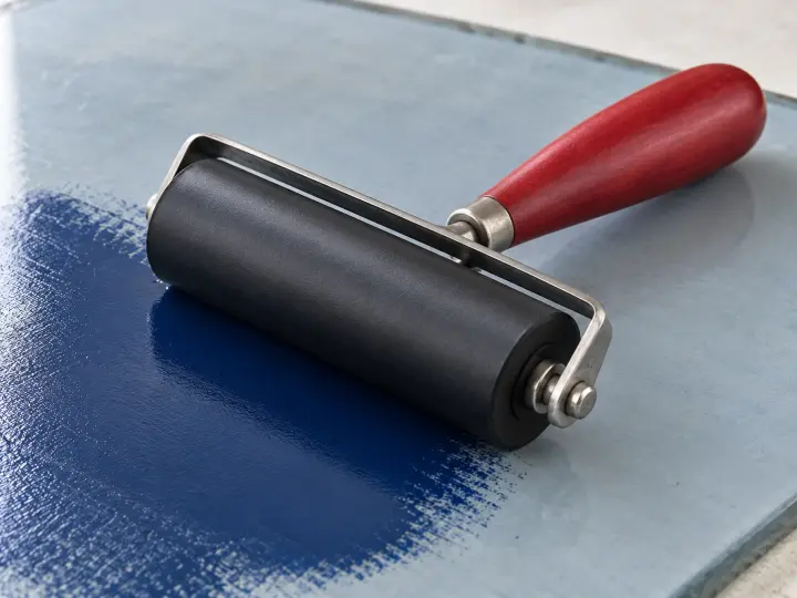 A black rubber printmaking brayer with a red handle resting beside blue ink on a pale slab.