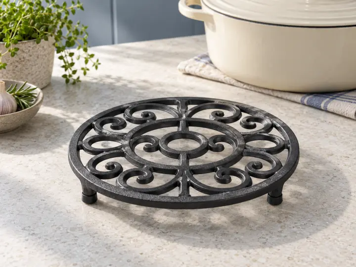 A round dark cast-iron trivet with open scrollwork and capped feet on a pale counter, with a cream casserole behind it.