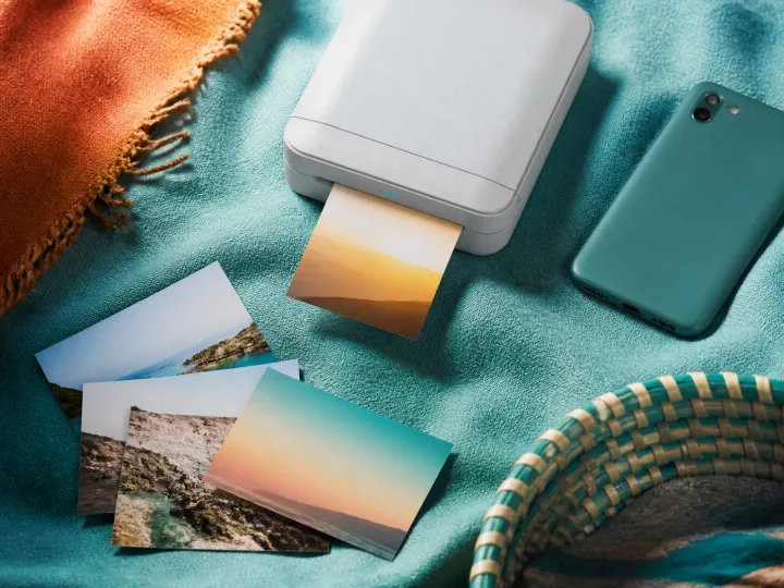 A compact photo printer producing a small landscape print beside several photographs and a face-down phone.