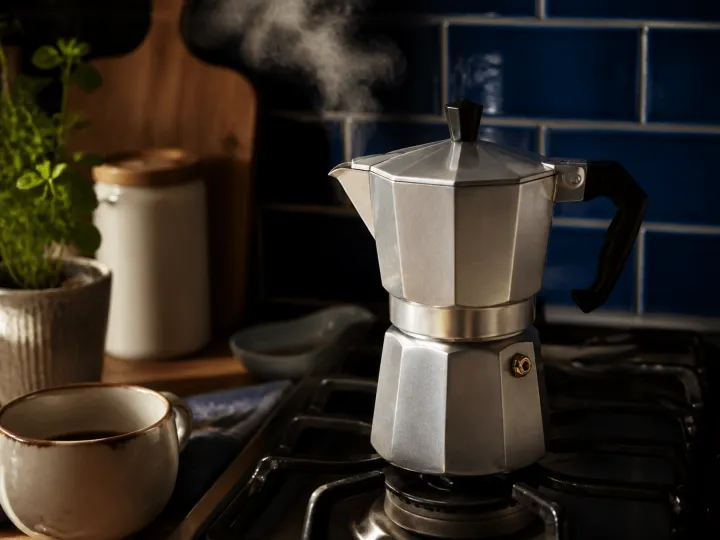 A silver moka pot steaming on a stovetop beside a ceramic coffee cup.